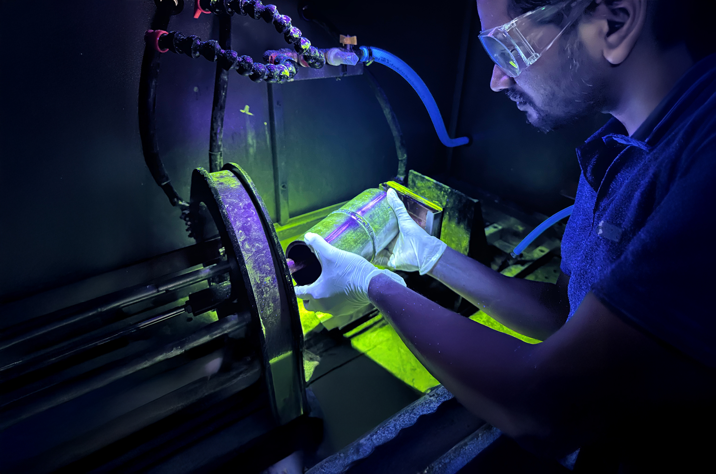 Technician wearing safety glasses and gloves performing a fluorescent magnetic particle inspection under ultraviolet (UV) light, examining a cylindrical metal component for surface cracks or defects.