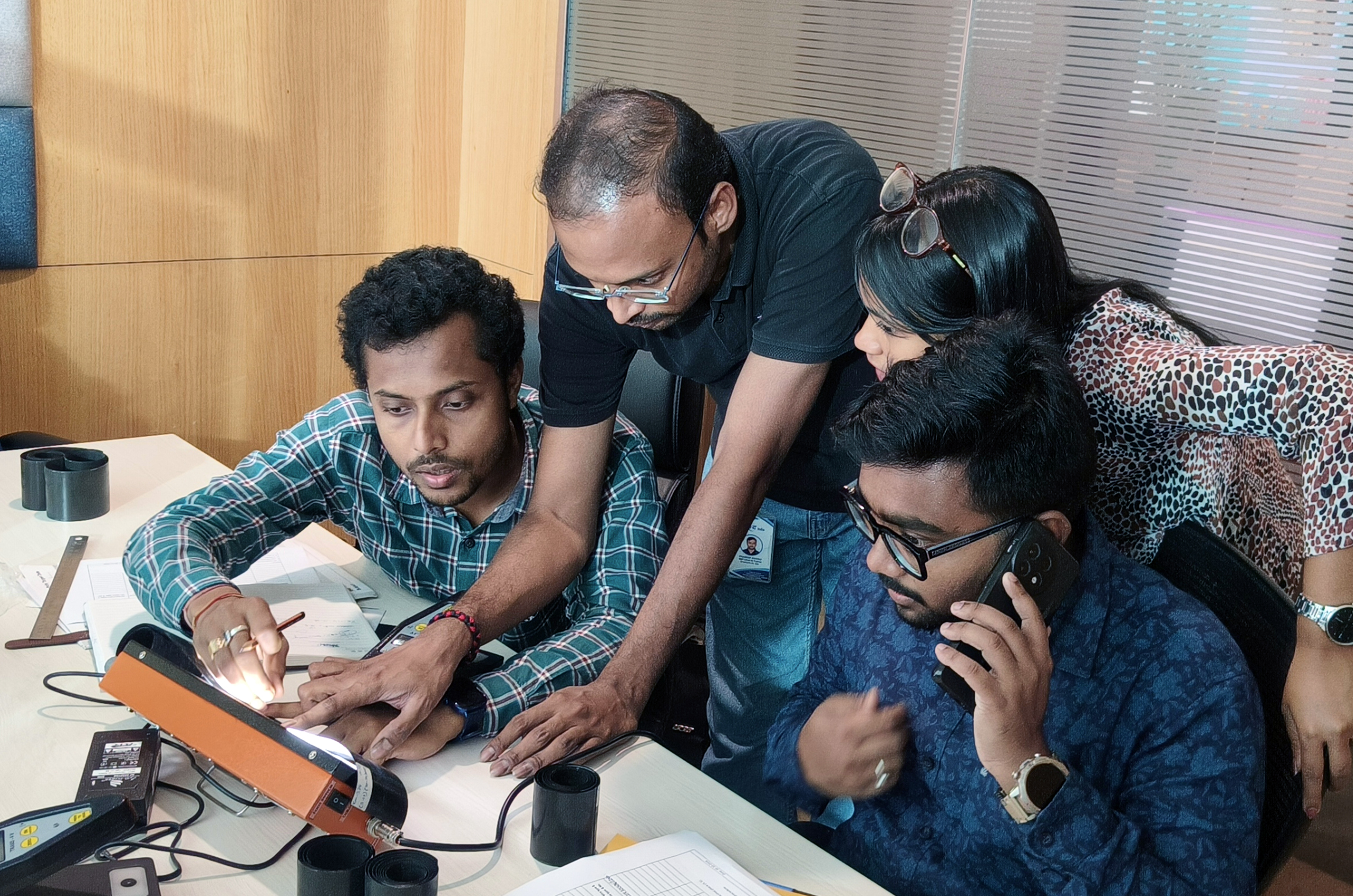 Group of four people collaborating at a desk, examining a sample while discussing results, with one team member on a phone call.