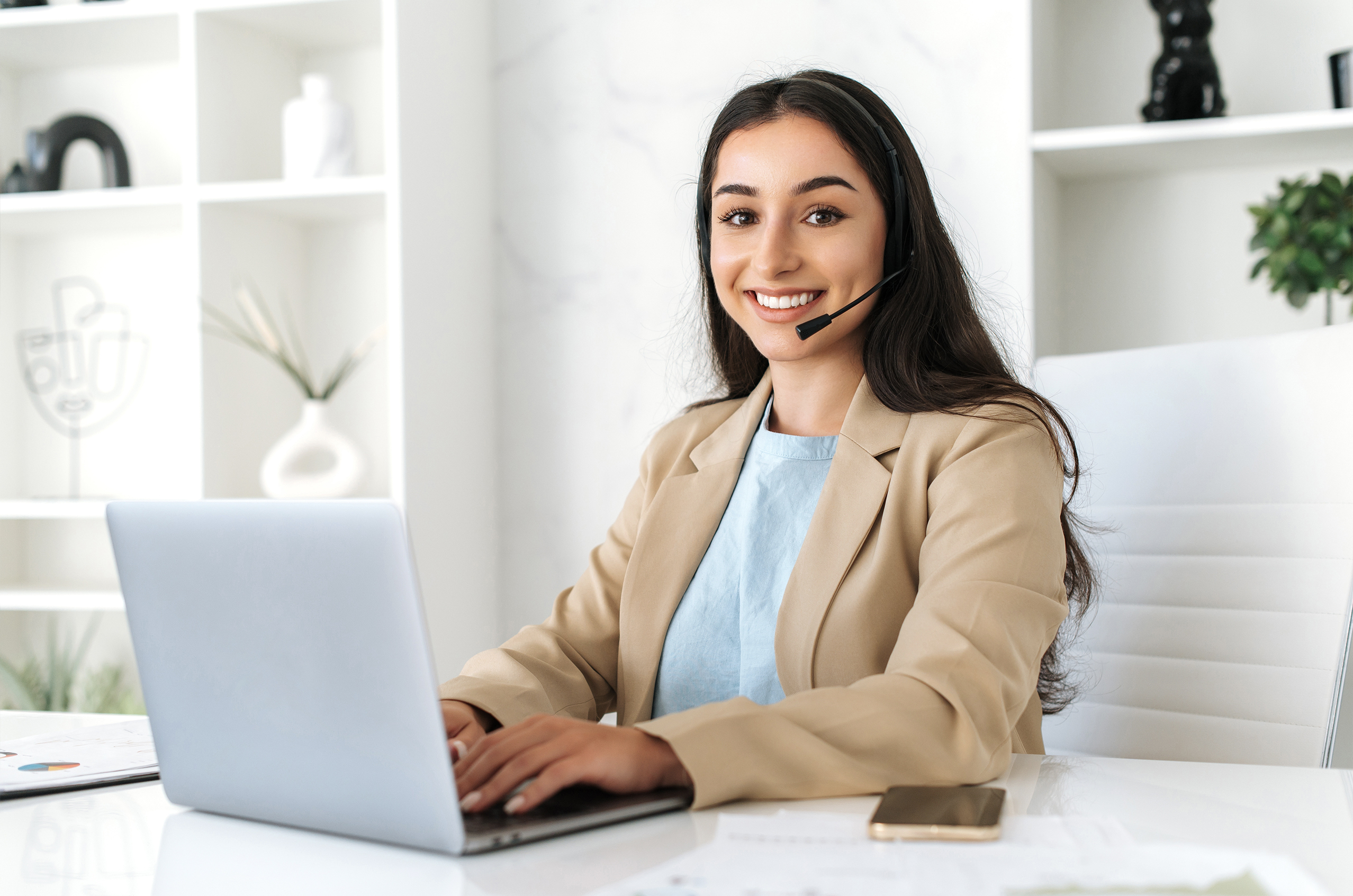 Smiling woman wearing a headset, working on a laptop at a desk in a modern office setting, providing customer support or online assistance.