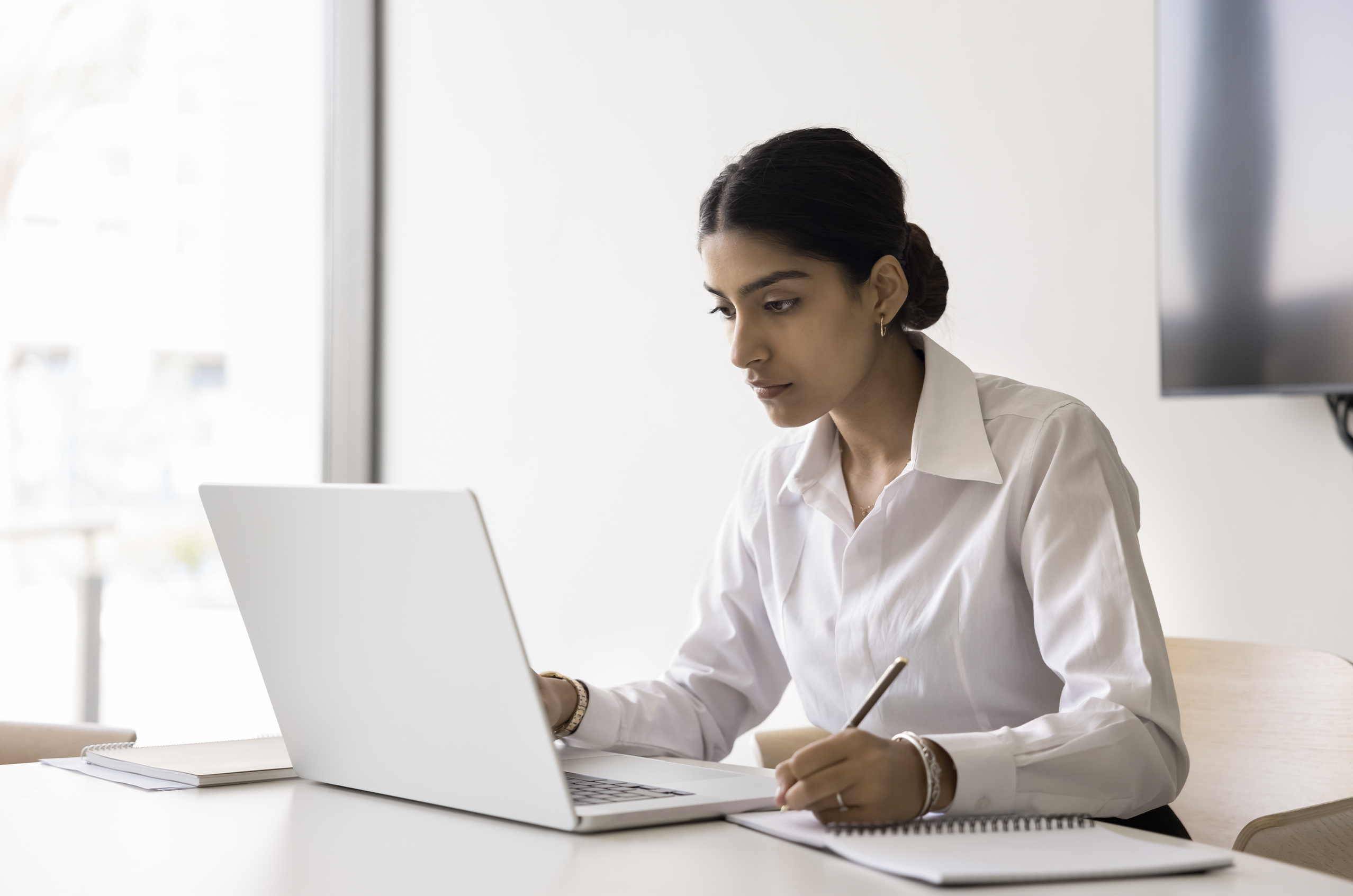 Woman in a white shirt working on a laptop while taking notes in a notebook, seated at a bright office desk near a window.